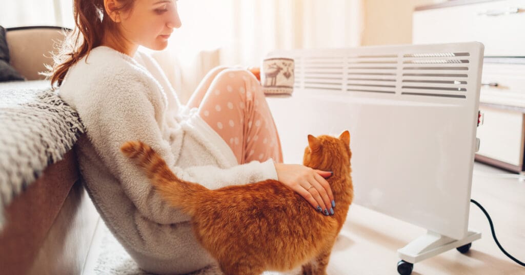 Lady keeping warm by radiator with cat and a cup of coffee during winter